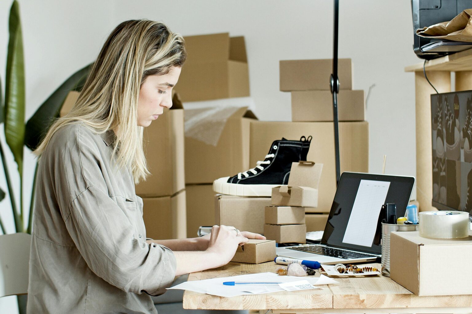 Comment se lancer dans la vente en ligne avec un investissement minimal A woman packing boxes for her online store, surrounded by packaging materials and a laptop.
