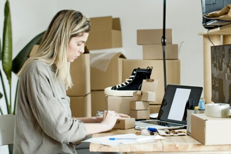 Comment se lancer dans la vente en ligne avec un investissement minimal A woman packing boxes for her online store, surrounded by packaging materials and a laptop.