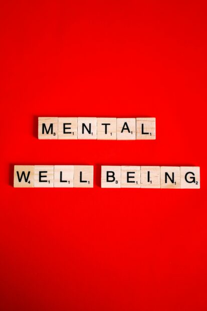 Souriez, vous êtes exploité·e : la psychologie positive est devenue une industrie à 6 800 milliards Scrabble tiles spelling 'mental well-being' on a red background, symbolizing mental health.