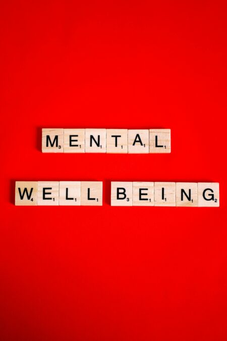 Souriez, vous êtes exploité·e : la psychologie positive est devenue une industrie à 6 800 milliards Scrabble tiles spelling 'mental well-being' on a red background, symbolizing mental health.