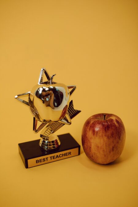 Cadeau maîtresse fin d’année 2026 : les vraies bonnes idées (et les pièges à éviter absolument) Award trophy and red apple set against a warm yellow backdrop symbolizing academic excellence.