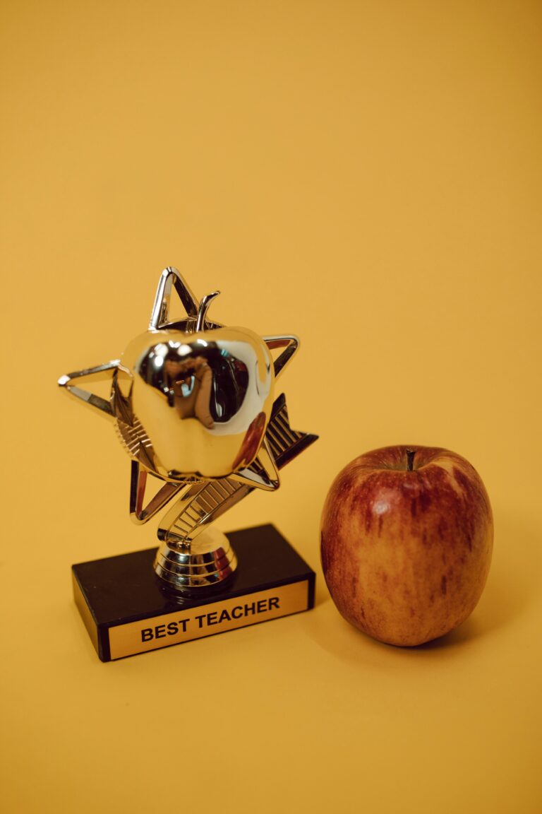 Cadeau maîtresse fin d’année 2026 : les vraies bonnes idées (et les pièges à éviter absolument) Award trophy and red apple set against a warm yellow backdrop symbolizing academic excellence.