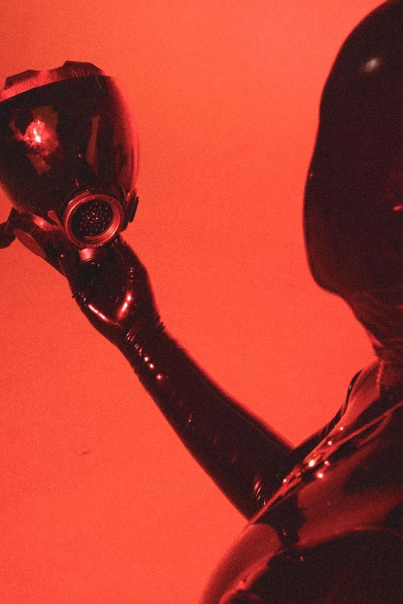A person in a latex suit holding a gas mask against a red background.