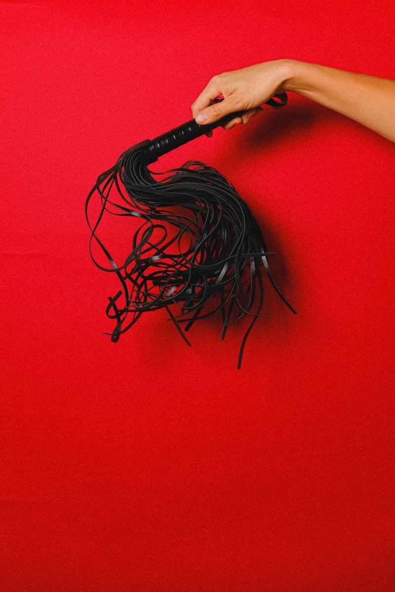 Black leather whip held by a hand on a vibrant red backdrop, symbolizing passion and sensuality.