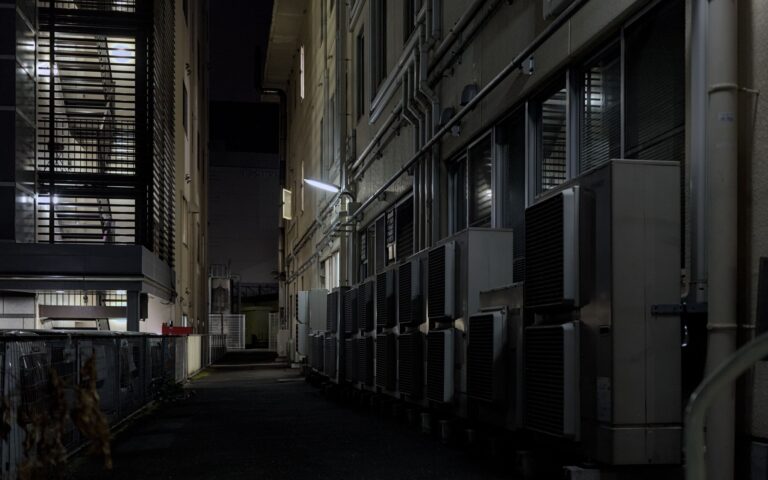 Poser une clim en pleine canicule : entre 1 500 et 15 000 €, et bonne chance pour trouver un artisan A dimly lit urban alley in Kyoto featuring air conditioning units, captured at night.