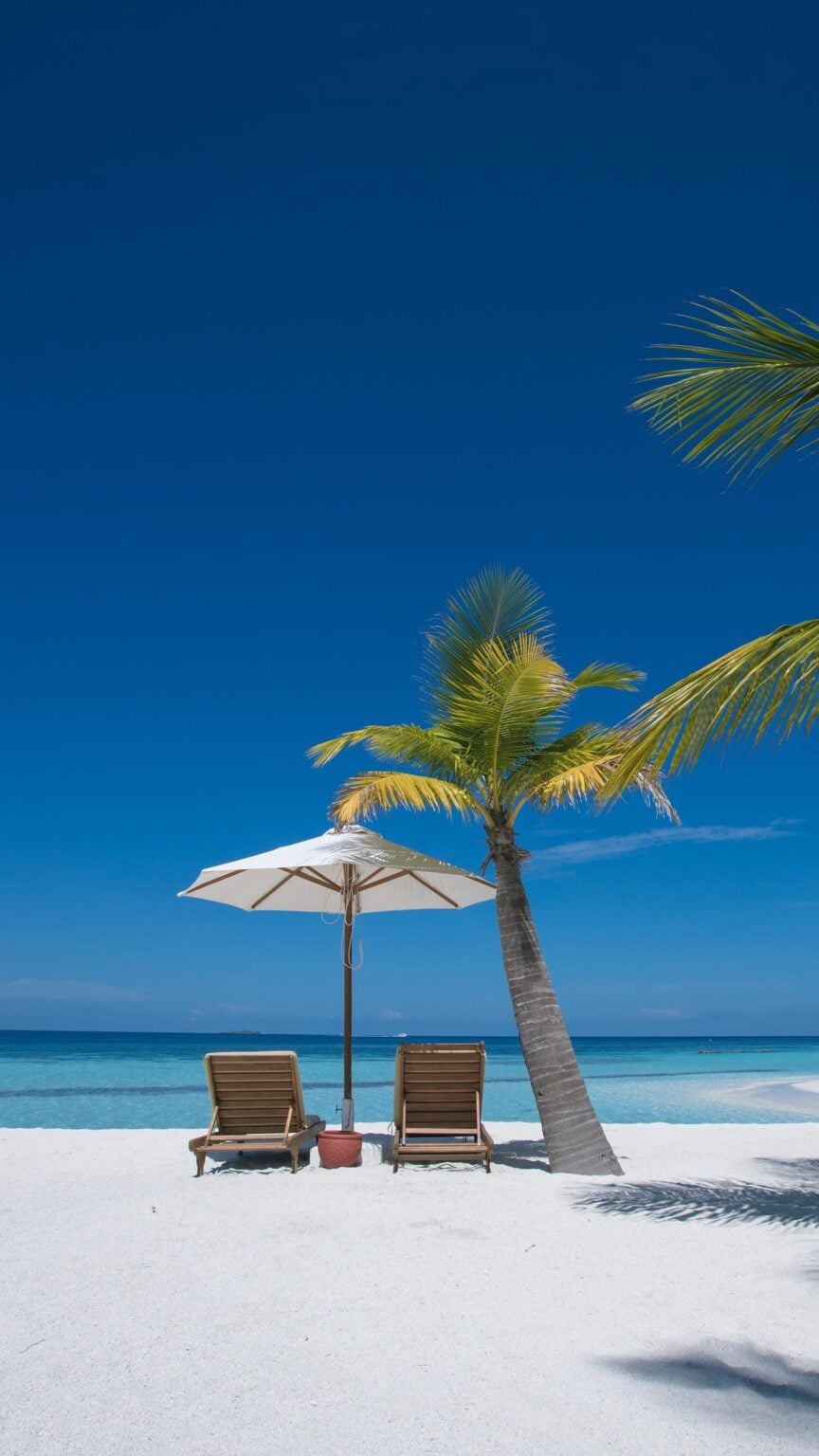 Vacances pas chères été 2026 : les pays où partir en juillet-août sans exploser votre budget Idyllic beachfront scene with two chairs under an umbrella by a palm tree on white sand.