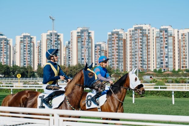 Quinté+ dans l’ordre : une chance sur 1 million et encore, c’est quand il y a peu de chevaux Jockeys on racehorses at a racetrack with a city skyline, combining sport and architecture.