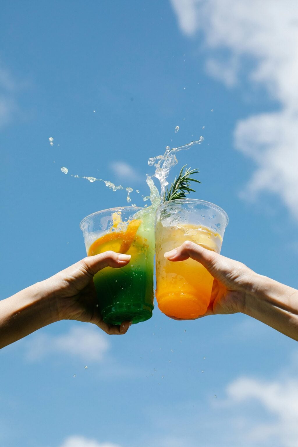 L’été et les cocktails : un duo incontournable Two colorful cocktails clinked against a bright blue sky, celebrating summer in style.