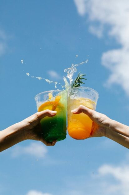 L’été et les cocktails : un duo incontournable Two colorful cocktails clinked against a bright blue sky, celebrating summer in style.