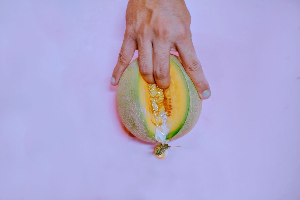 A hand gently touches a ripe melon on a pastel pink backdrop, creating a vivid and conceptual image.