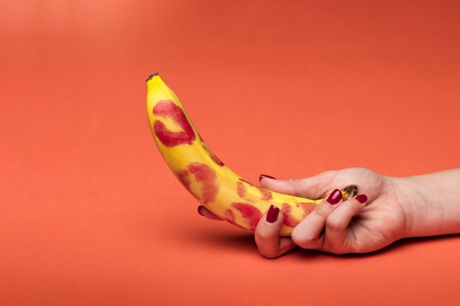 Meilleurs sites libertins 2026 : classement pour des rencontres réelles A feminine hand holds a banana covered in lipstick marks on a vibrant red background.