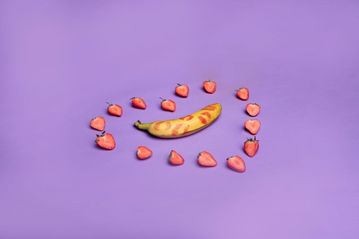 Creative studio photo of a banana with lipstick marks surrounded by strawberries on a purple background.