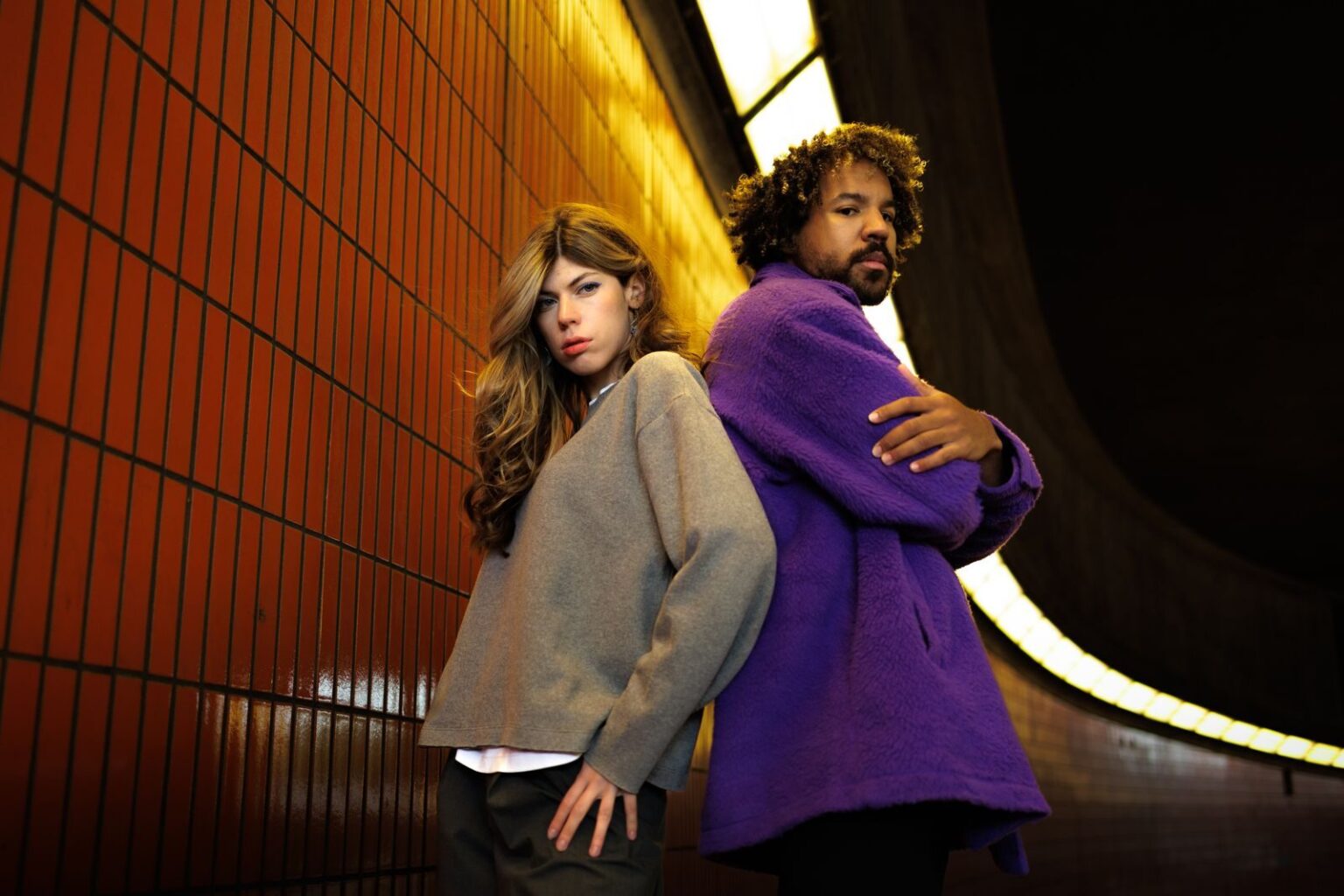 Aubergine, violet royal et blanc fantôme : les couleurs qui vont dicter nos garde-robes jusqu’à Noël 2026 Stylish individuals pose against a tiled wall in a dimly lit urban tunnel, expressing modern fashion.