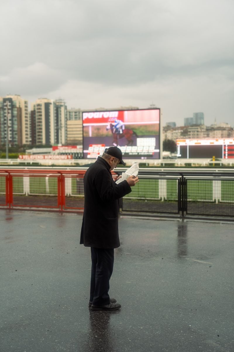 Comment les bookmakers établissent leurs cotes : derrière les chiffres, une mécanique bien huilée An elderly man reads a newspaper at a rainy horse race track with city skyline in the background.