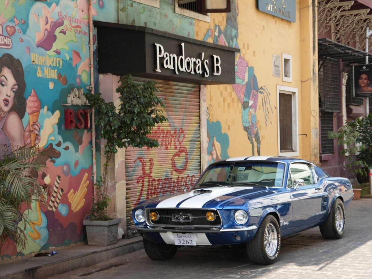 A classic blue Ford Mustang parked beside a vibrant mural in Mumbai, India, showcasing street art.