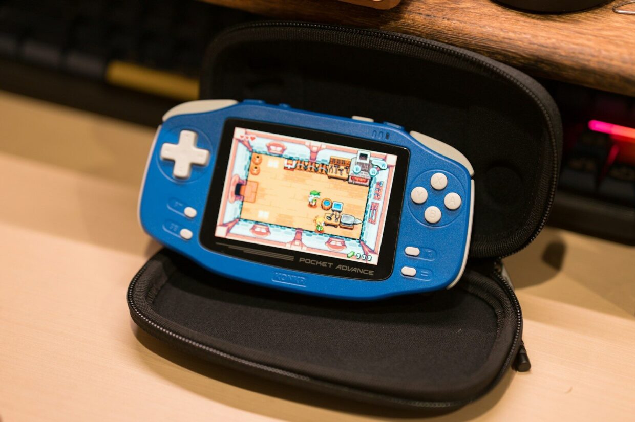 A blue handheld game console displaying a pixelated interior scene in a black case