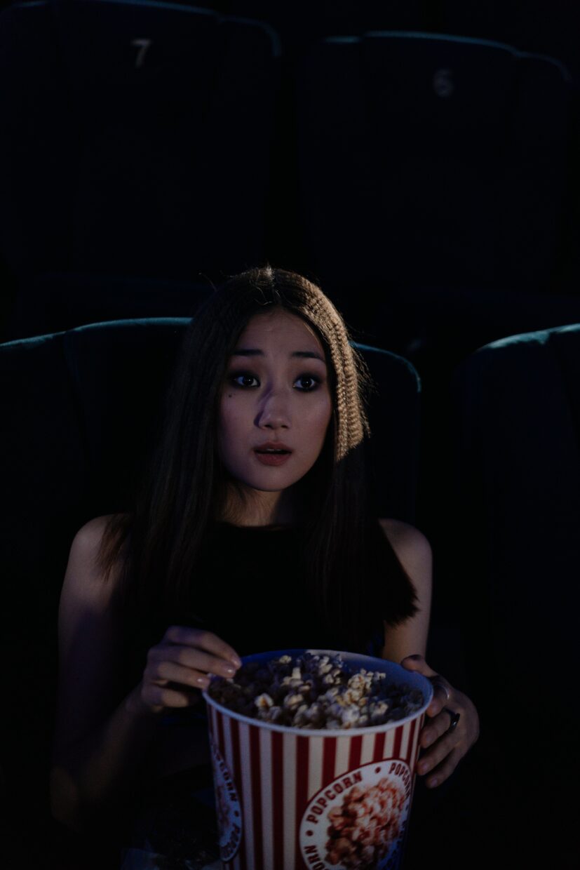 Asian woman holding popcorn looking surprised in a dark cinema.