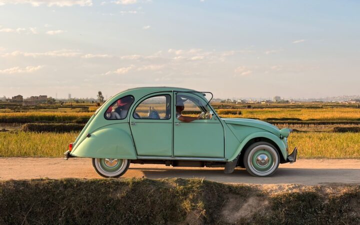 Classic mint-green car driving down a rural road in Madagascar under a sunny sky with scenic fields.