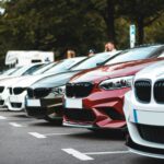 A row of parked cars including white, olive, and red sedans in a lot